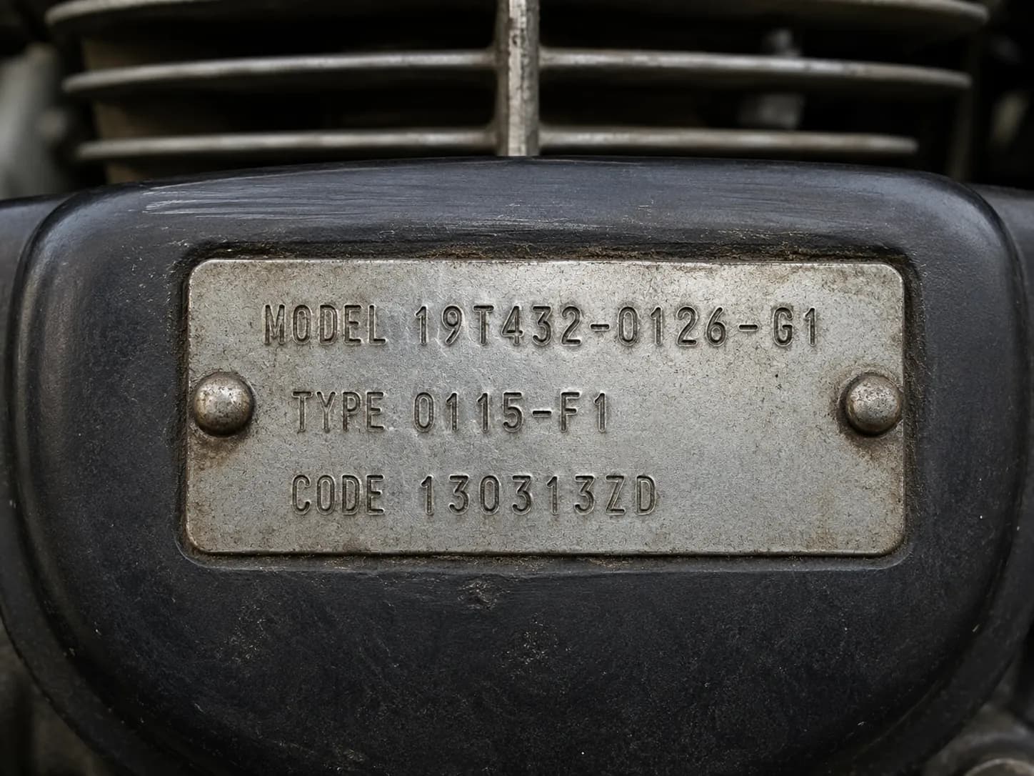 Close-up of a small engine identification plate stamped with model, type, and code numbers, riveted to a matte black engine shroud with cooling fins softly blurred in the background.
