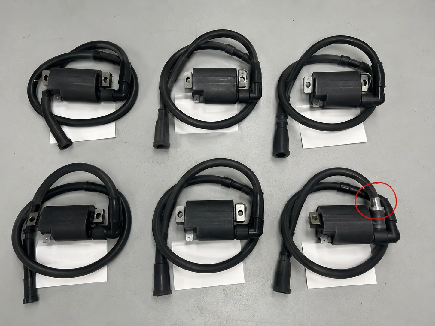 Six small engine ignition coil assemblies arranged for comparison, with one lower-right coil showing a threaded spark plug wire connection circled in red on a light gray workbench.