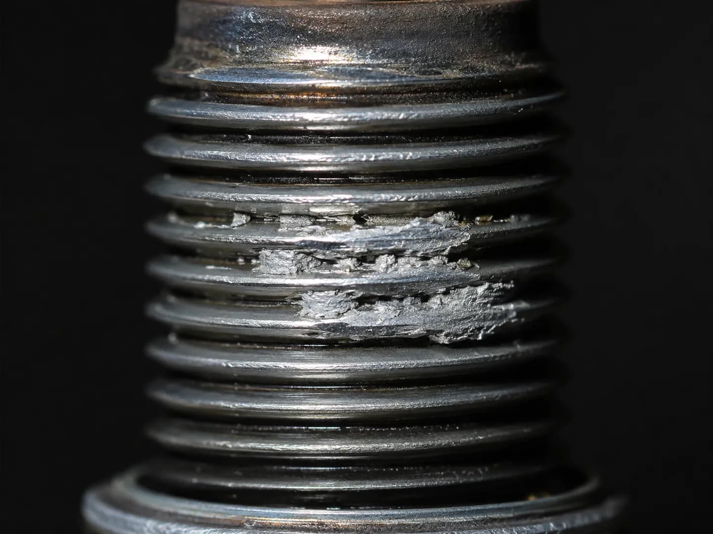 Extreme macro of a removed spark plug with light gray aluminum smeared into the threads, showing cylinder head thread transfer caused by seized or cross-threaded spark plug removal.