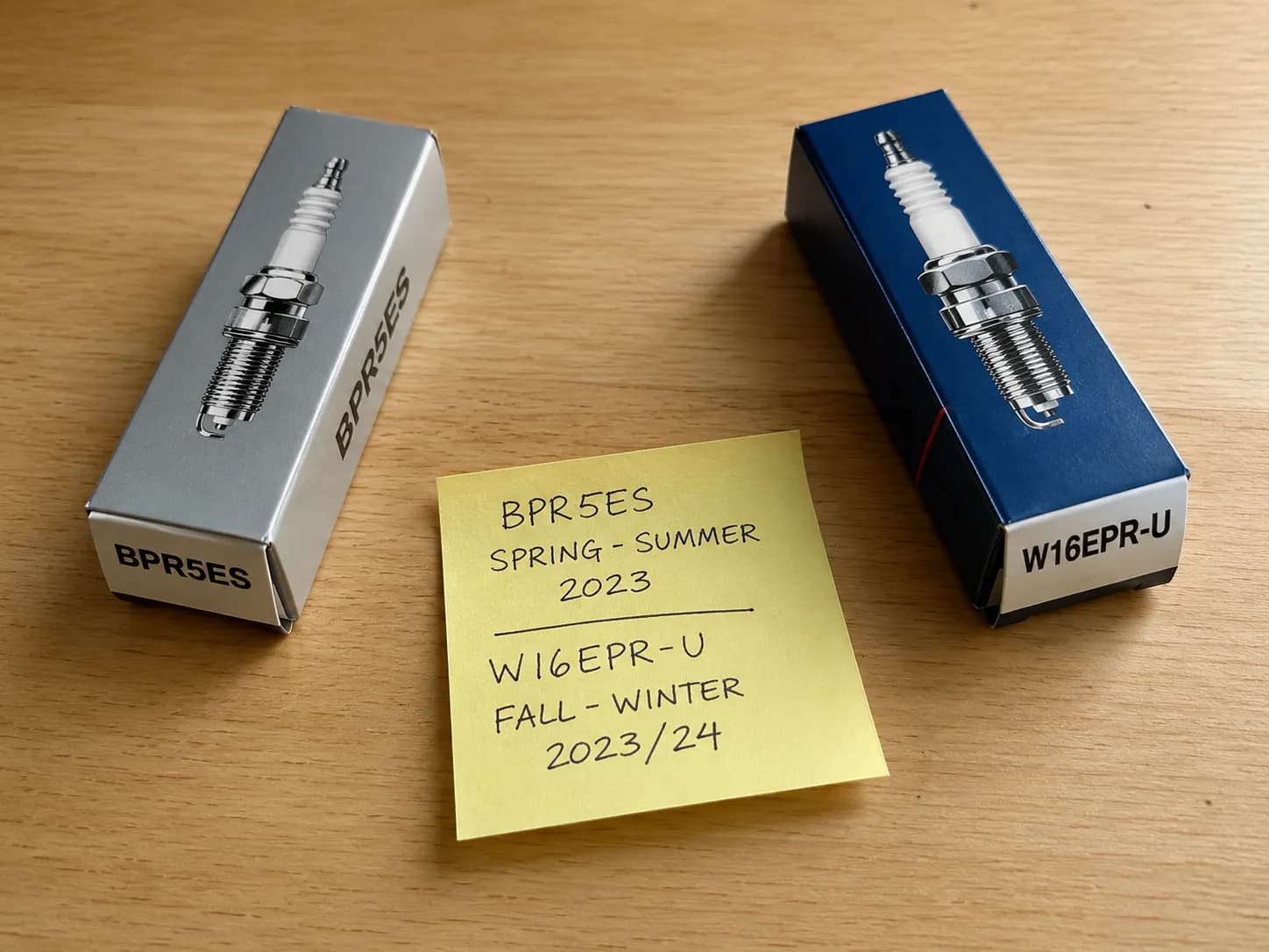 Overhead view of two spark plug boxes placed side by side on a wooden surface with a handwritten sticky note recording the season each equivalent plug was used for comparison.