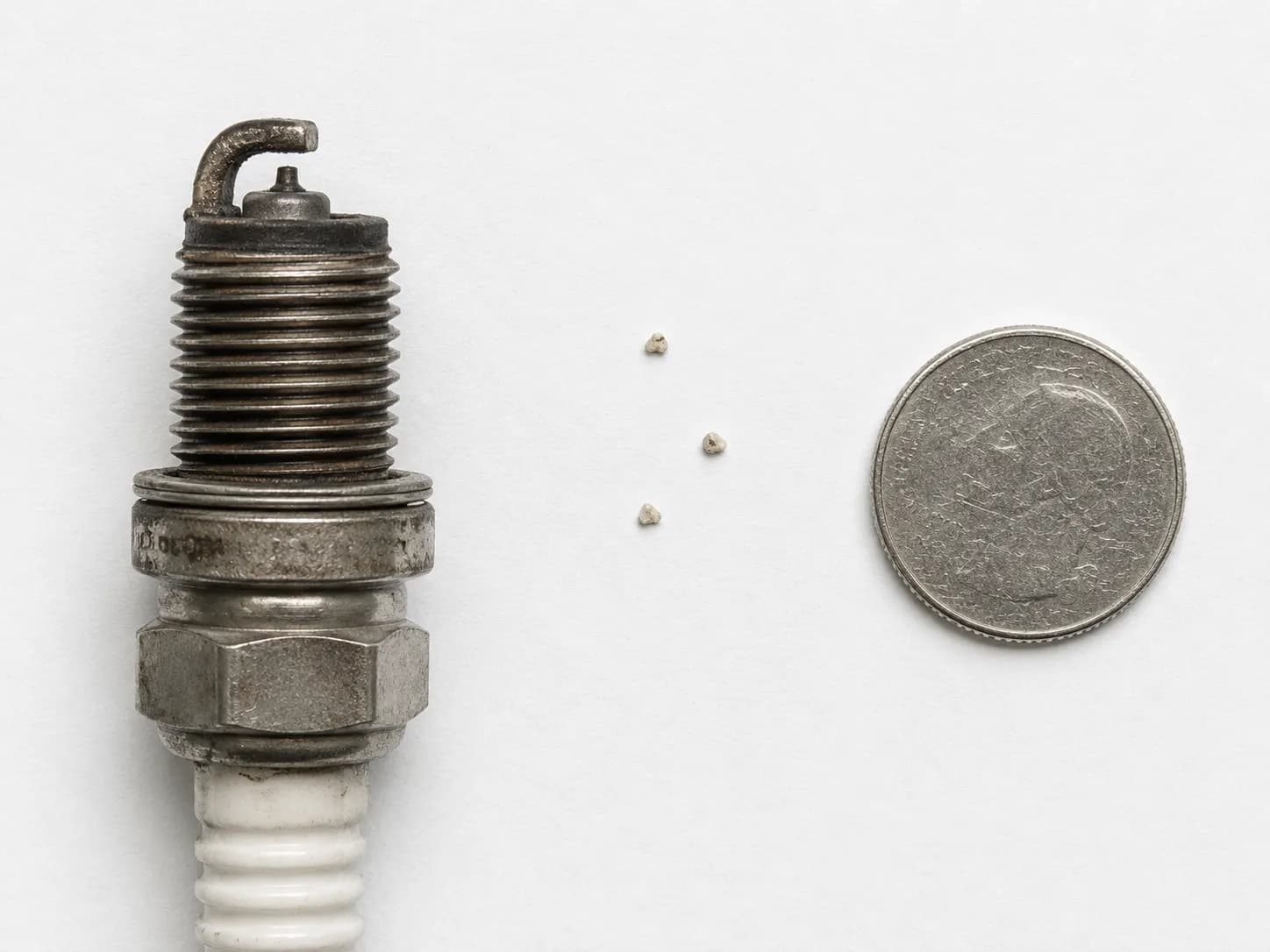 Macro view of three abrasive media particles next to a used spark plug and a coin, illustrating the small amount of blasting residue remaining after air cleaning.