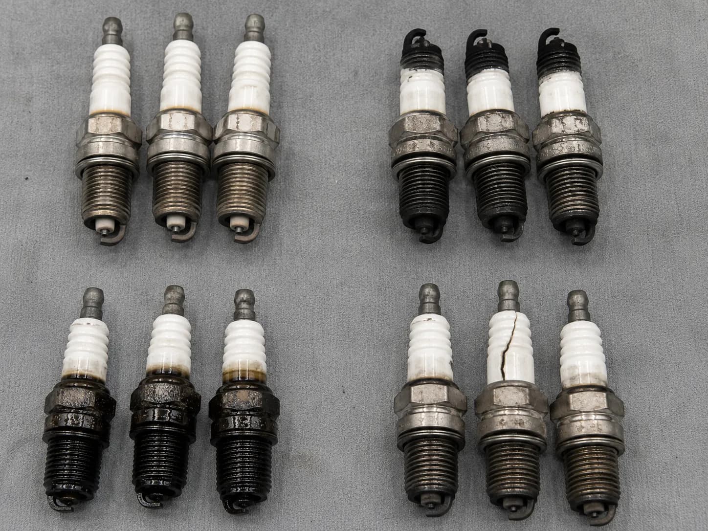 Groups of used lawn mower spark plugs organized by deposit type, including normal light tan, dry carbon fouling, wet oil fouling, and a cracked insulator for engine diagnosis and maintenance.