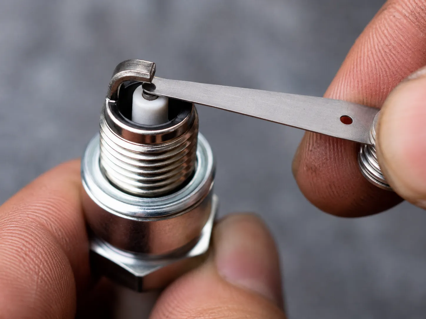 Feeler gauge checking the electrode gap on a new lawn mower spark plug, showing the ceramic insulator and metal electrodes in a close-up workshop view.