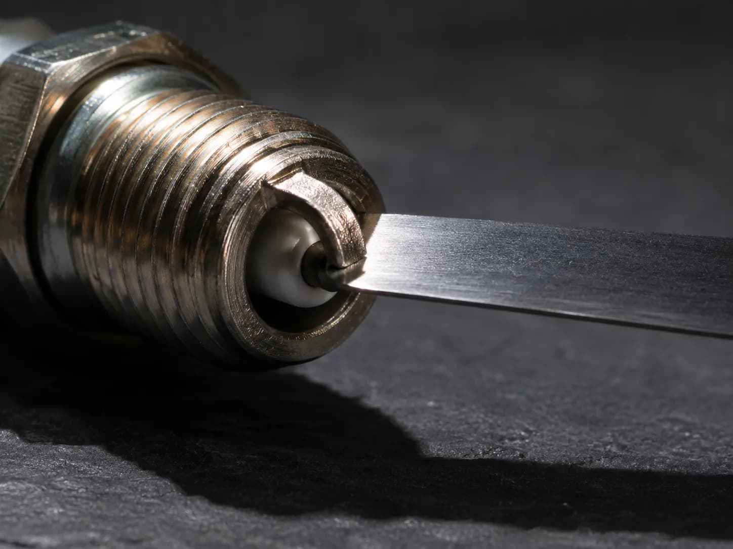 Extreme macro of a feeler gauge measuring the electrode gap on a new spark plug, illustrating a factory gap that is 0.005 inches wider than specification before installation.