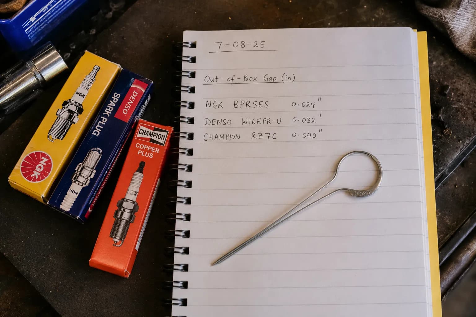 Overhead view of a notebook listing three spark plug gap measurements beside three spark plug boxes and a wire gap gauge on a garage workbench under warm task lighting.