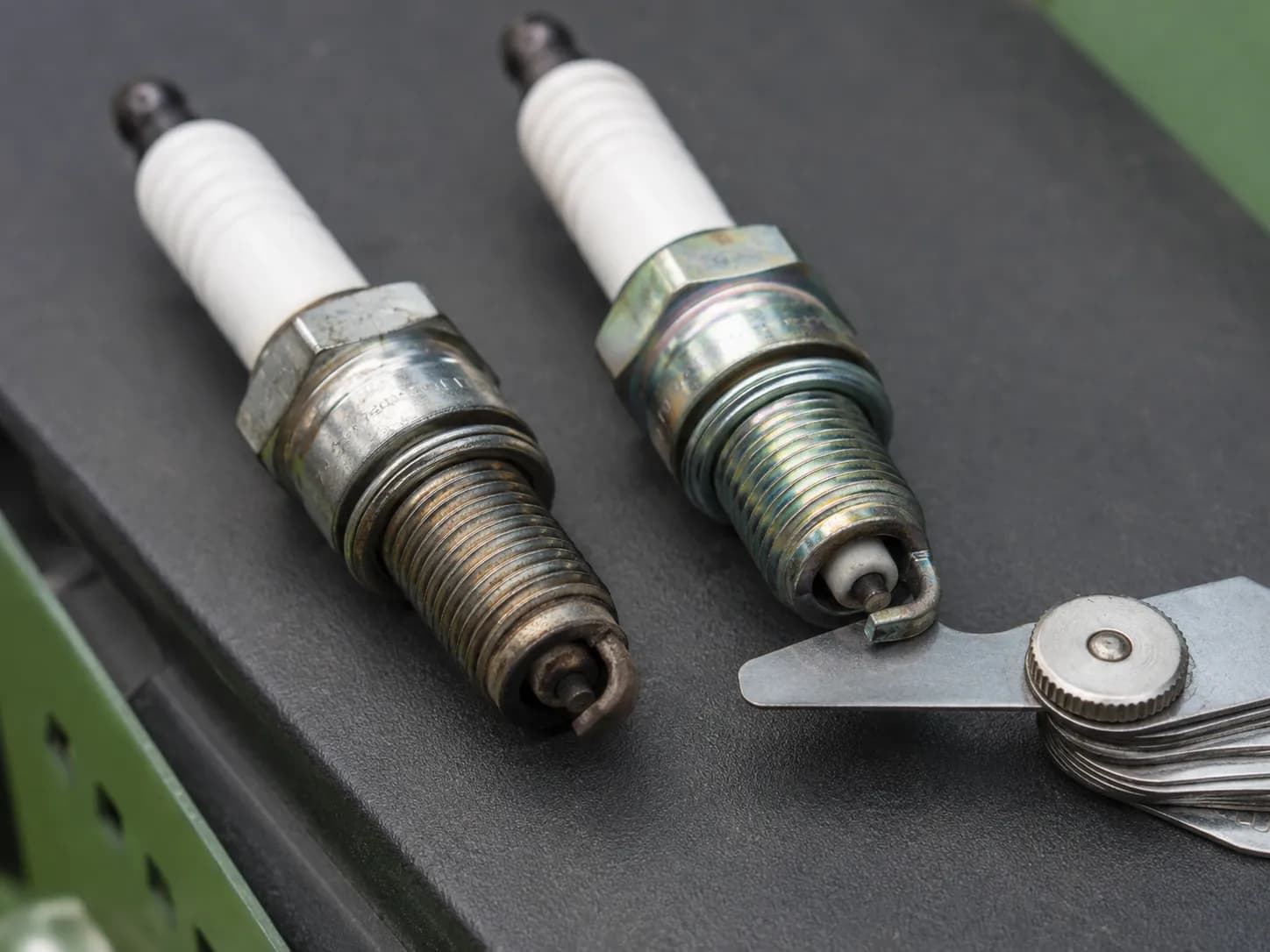 Two small-engine spark plugs, an RJ19LM and an RC12YC, lying side by side on a graphite workbench while a coin-style feeler gauge measures the electrode gap on the nearer plug.