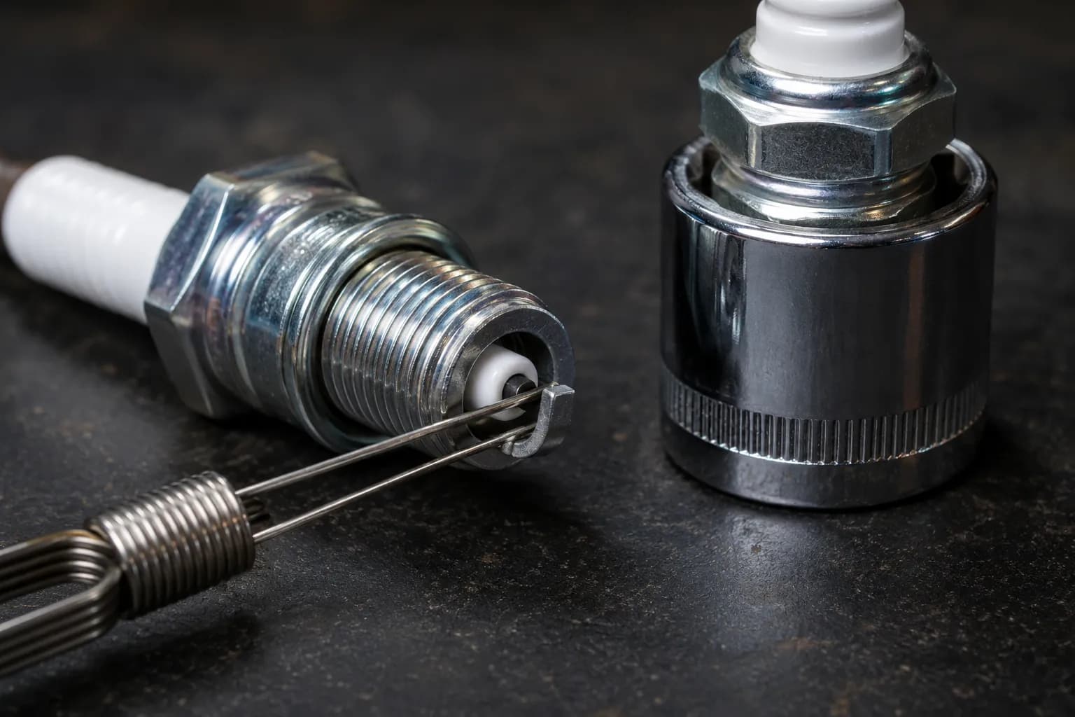 Wire feeler gauge checking the electrode gap on a clean BPR5ES spark plug beside a 13/16-inch spark plug socket on a dark workbench with sharp macro focus.