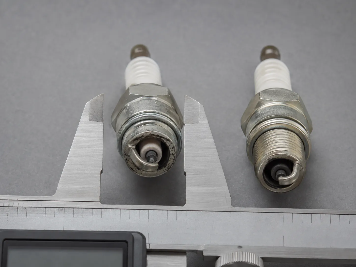 Two small engine spark plugs with a stainless digital caliper measuring the hex size of the nearer plug on a grey surface, comparing actual measurements against published spark plug charts.