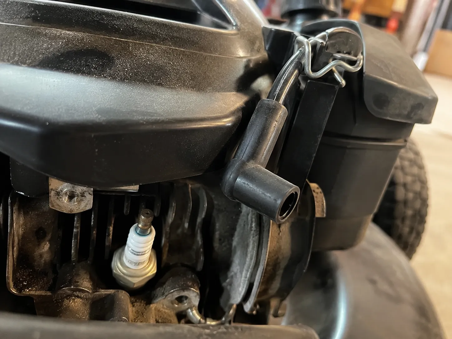 Black rubber spark plug lead removed from a small mower engine and clipped safely aside with a spring clamp before maintenance, leaving the spark plug exposed for safe servicing.