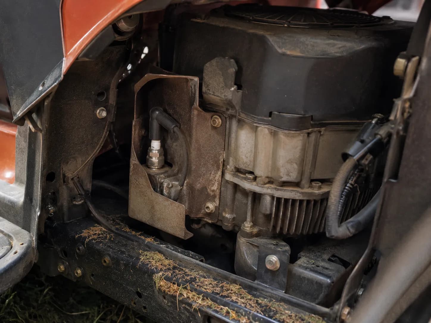 Side view inside a riding lawn mower engine bay showing the spark plug behind a metal heat shield with the black ignition boot attached and ignition lead running toward the coil.