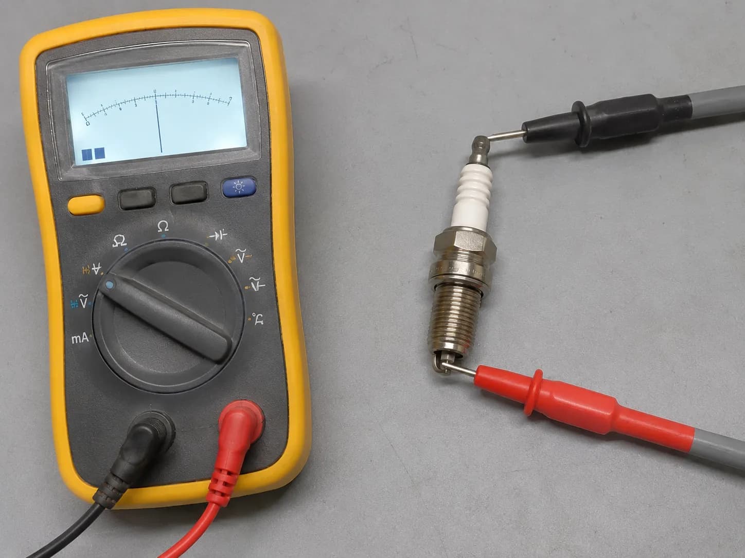 Digital multimeter measuring spark plug resistance with probes touching the terminal nut and center electrode during an ohms continuity test on a workbench.