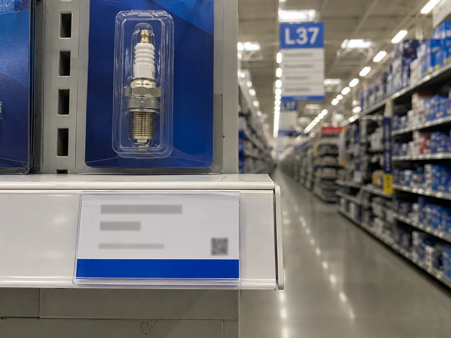 Close-up of a spark plug shelf price tag beneath a packaged small engine spark plug in a brightly lit retail aisle, with an L37 aisle marker softly blurred in the background.