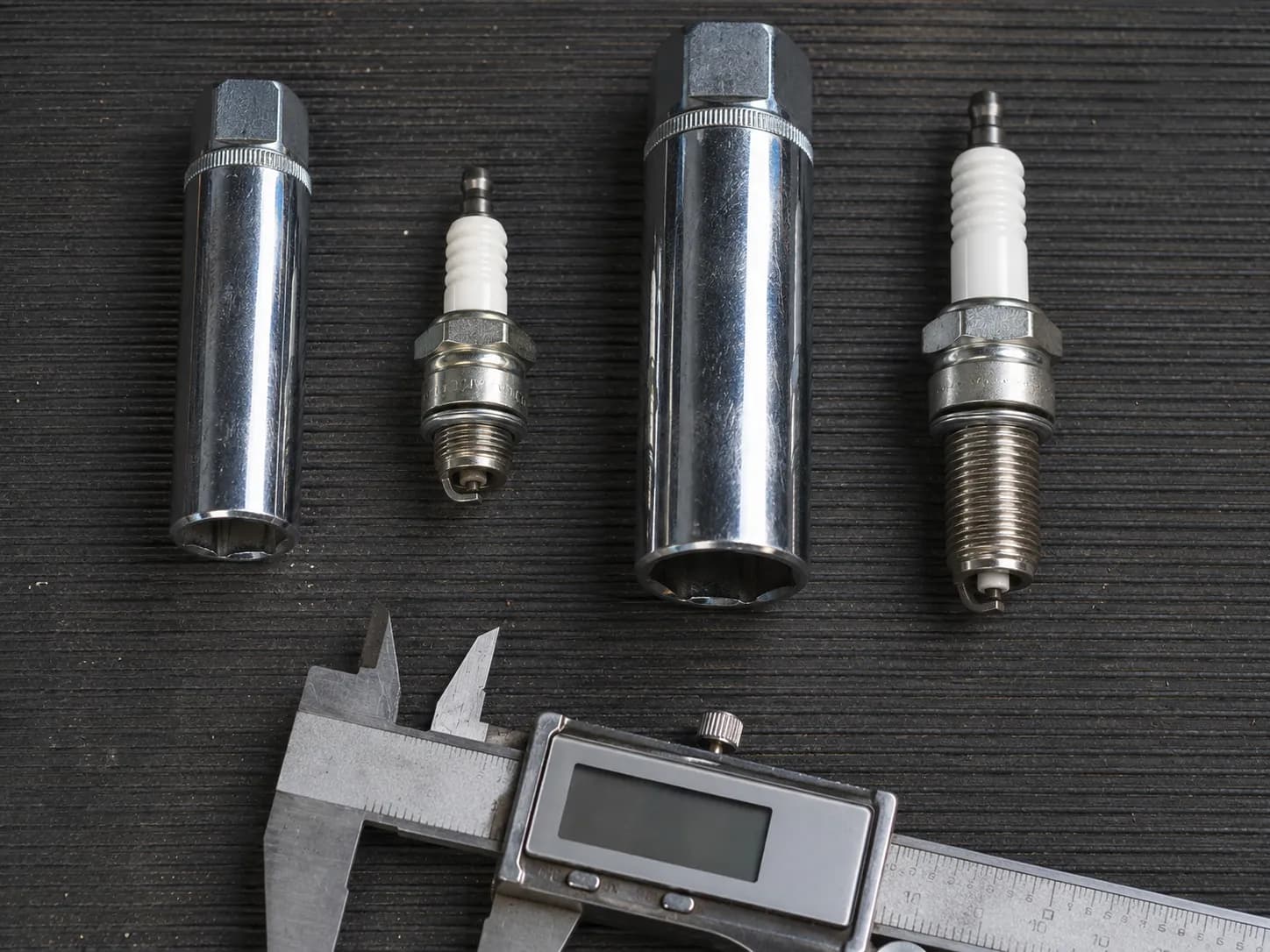 Two different size spark plug sockets paired with matching spark plugs and a digital caliper on a dark workshop bench.