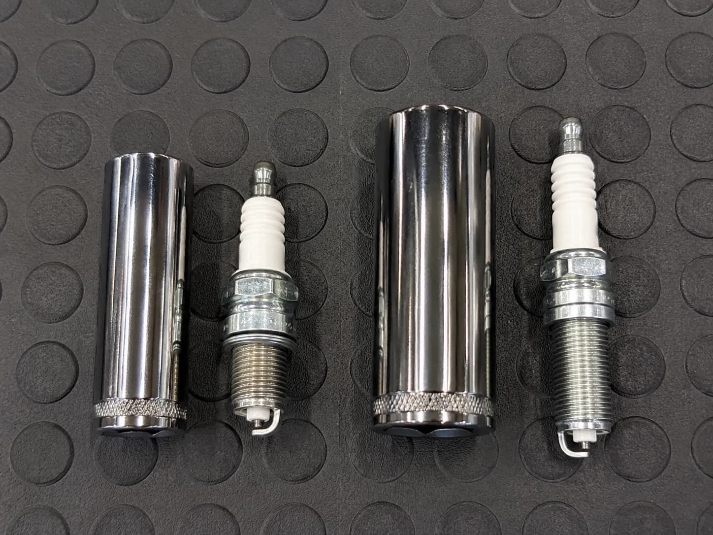5/8-inch and 13/16-inch spark plug sockets placed beside RJ19LM and RC12YC spark plugs, showing which socket fits each plug on a dark rubber work mat.