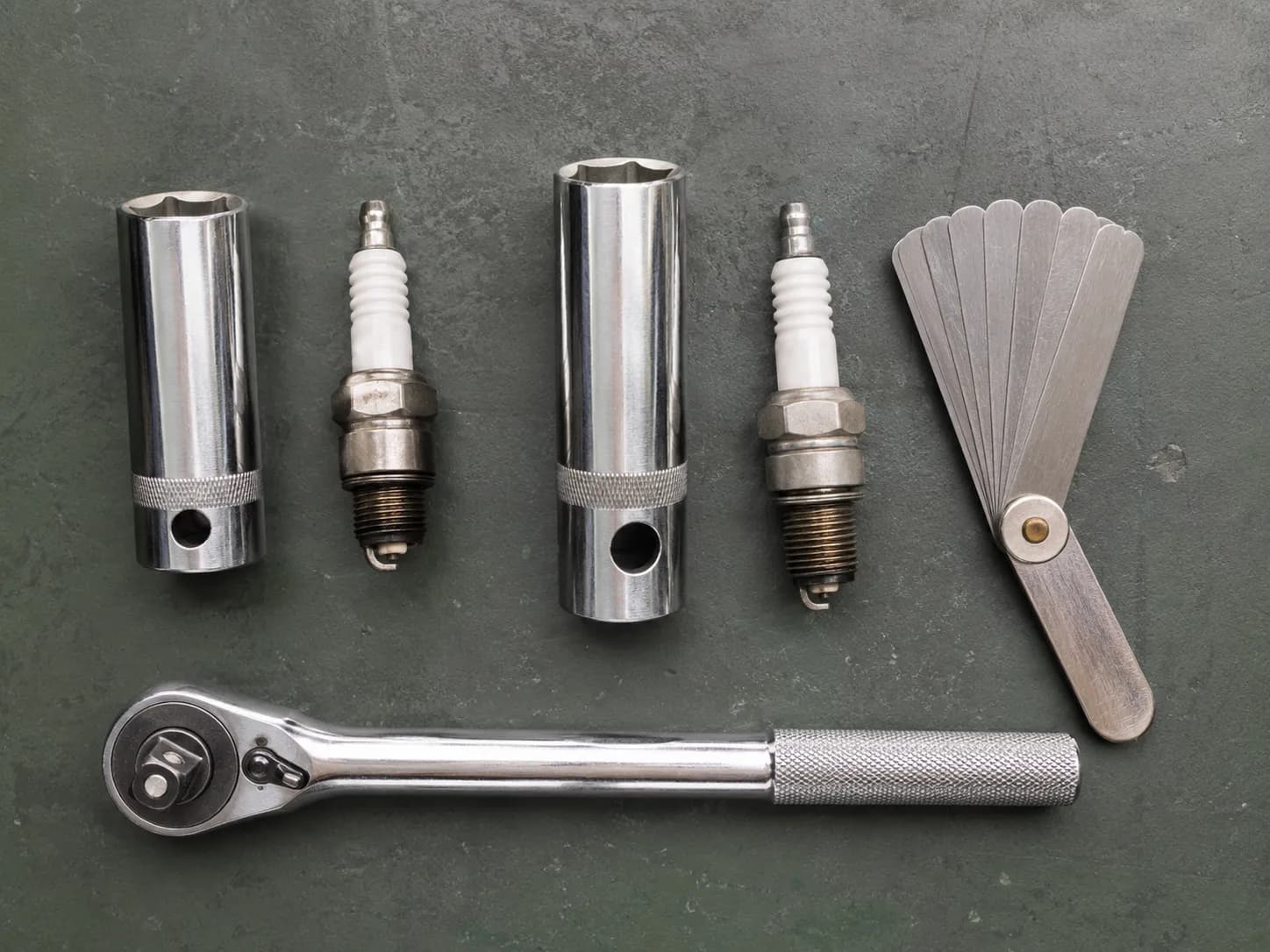 Top-down view of 5/8-inch and 13/16-inch spark-plug sockets placed beside the small-engine spark plugs they fit, with a feeler gauge and 3/8-inch ratchet on a graphite workbench.