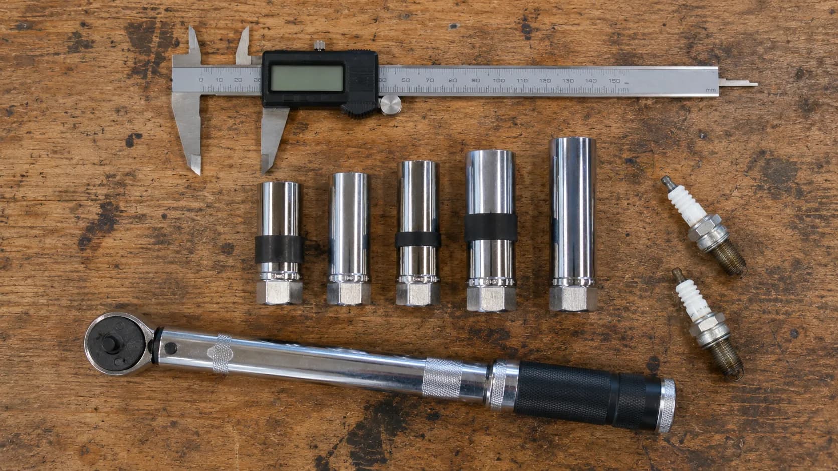 Overhead view of five spark plug sockets selected from testing, alongside a digital caliper, 3/8-inch torque wrench, and two used spark plugs on a wooden workbench.