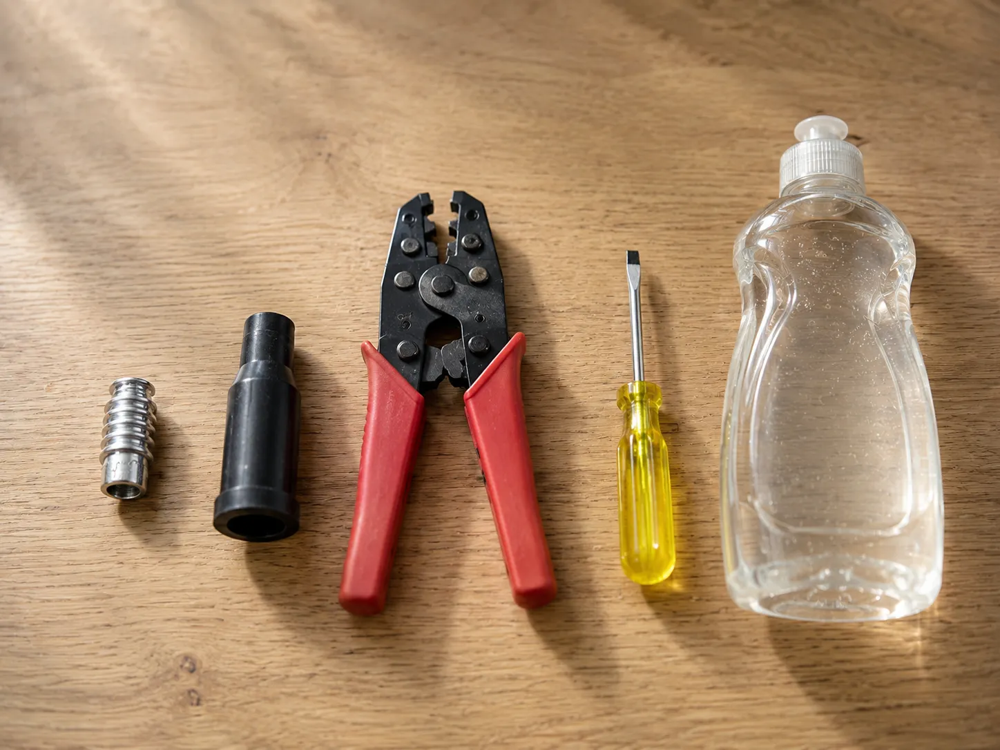 Flat lay of spark plug wire repair tools including a spark plug terminal, rubber boot, crimping pliers, flat-head screwdriver, and clear dish soap bottle on a wooden surface.