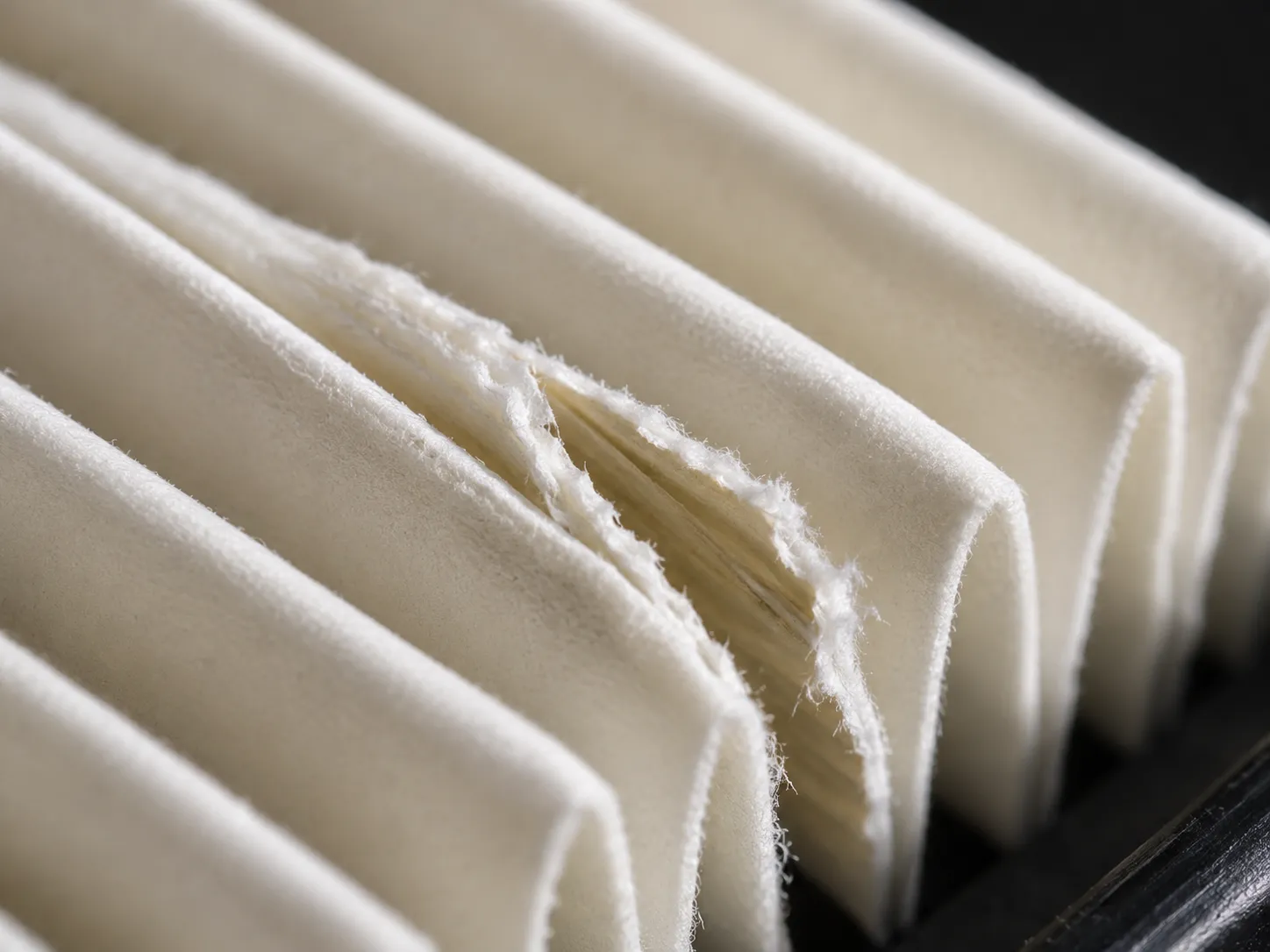 Macro close-up of a split pleated paper lawn mower air filter seam beside an intact pleat, highlighting torn fibers and damage compared with an undamaged filter fold.