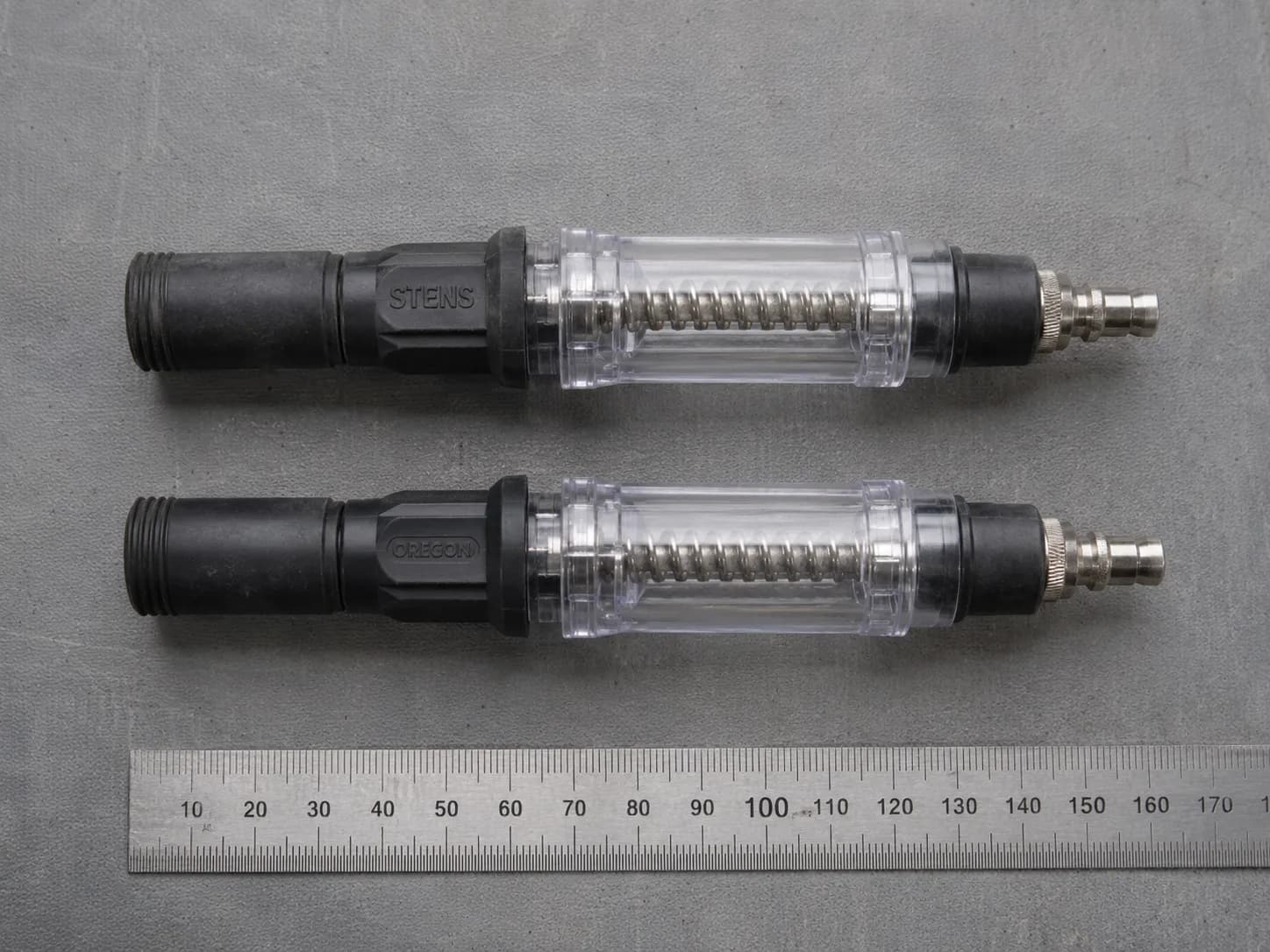 Stens and Oregon inline spark testers shown side by side on a workbench with a steel ruler for scale, highlighting their nearly identical design apart from the moulded lettering.