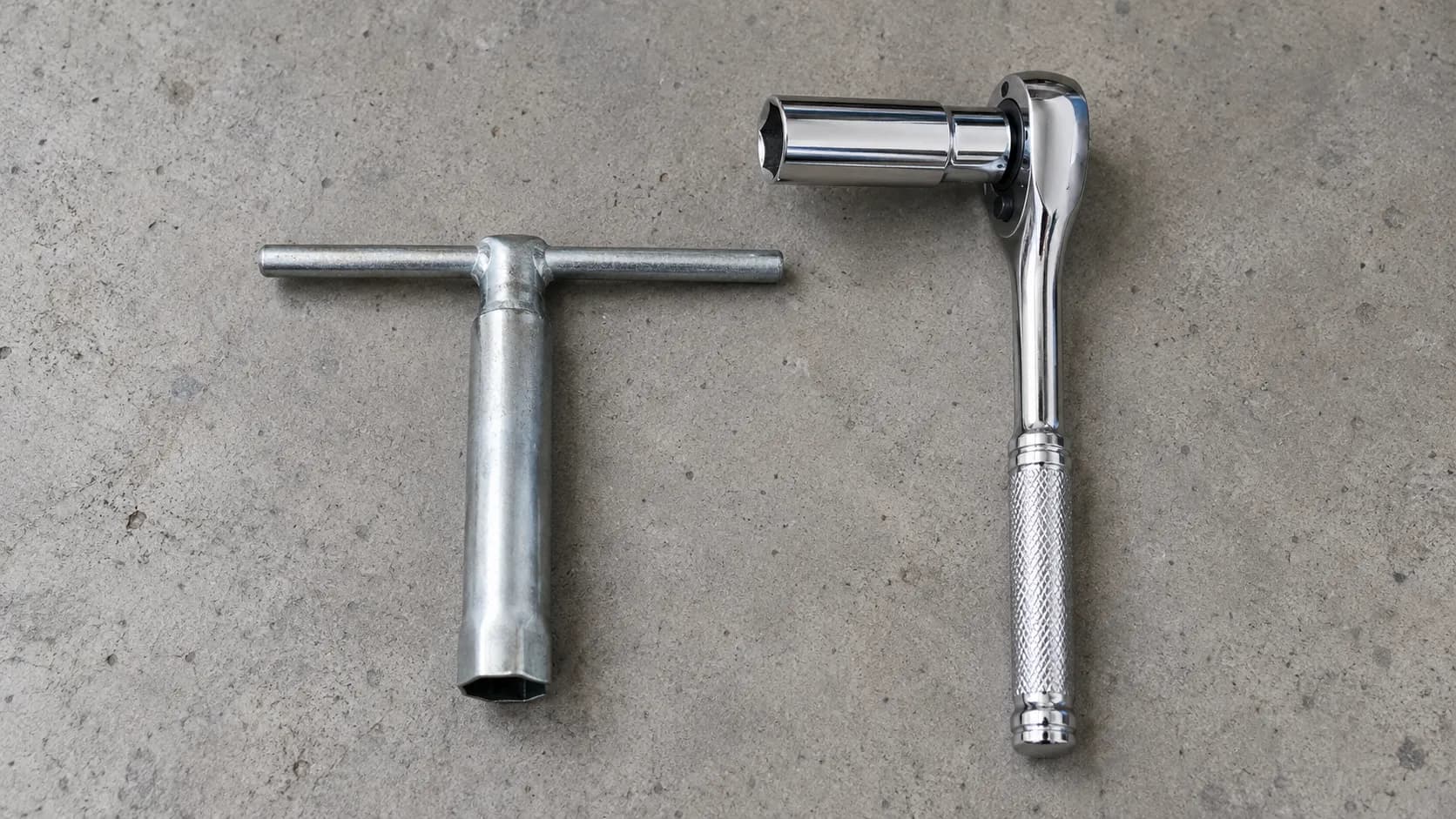Stamped steel T-handle spark plug wrench beside a 3/8-inch ratchet with a spark plug socket, comparing the basic tool supplied with small engines to a standard ratchet setup.