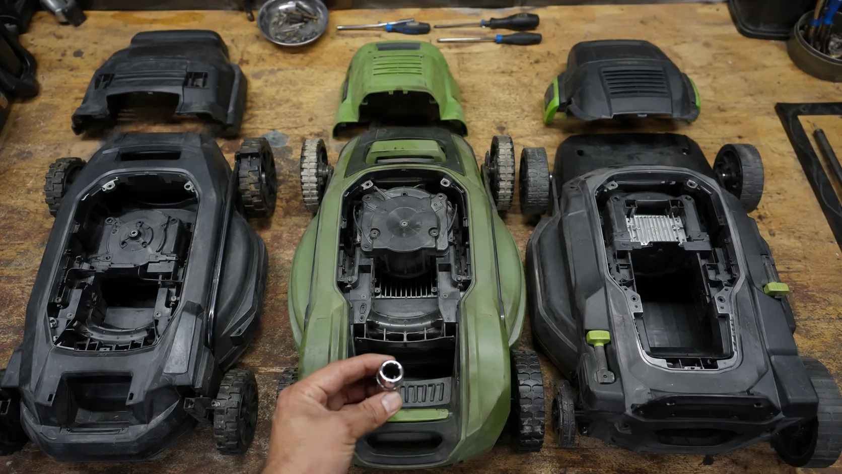 Overhead view of three cordless electric lawn mowers opened on a workbench with motor housings removed, while a hand holds a spark plug socket to show there are no spark plugs, air filters, dipsticks.
