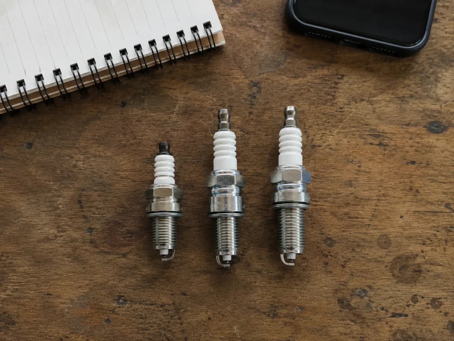Three lawn mower spark plugs arranged on a wooden workbench, including one shorter plug beside two longer ones, with a notebook and phone in the background for size comparison.