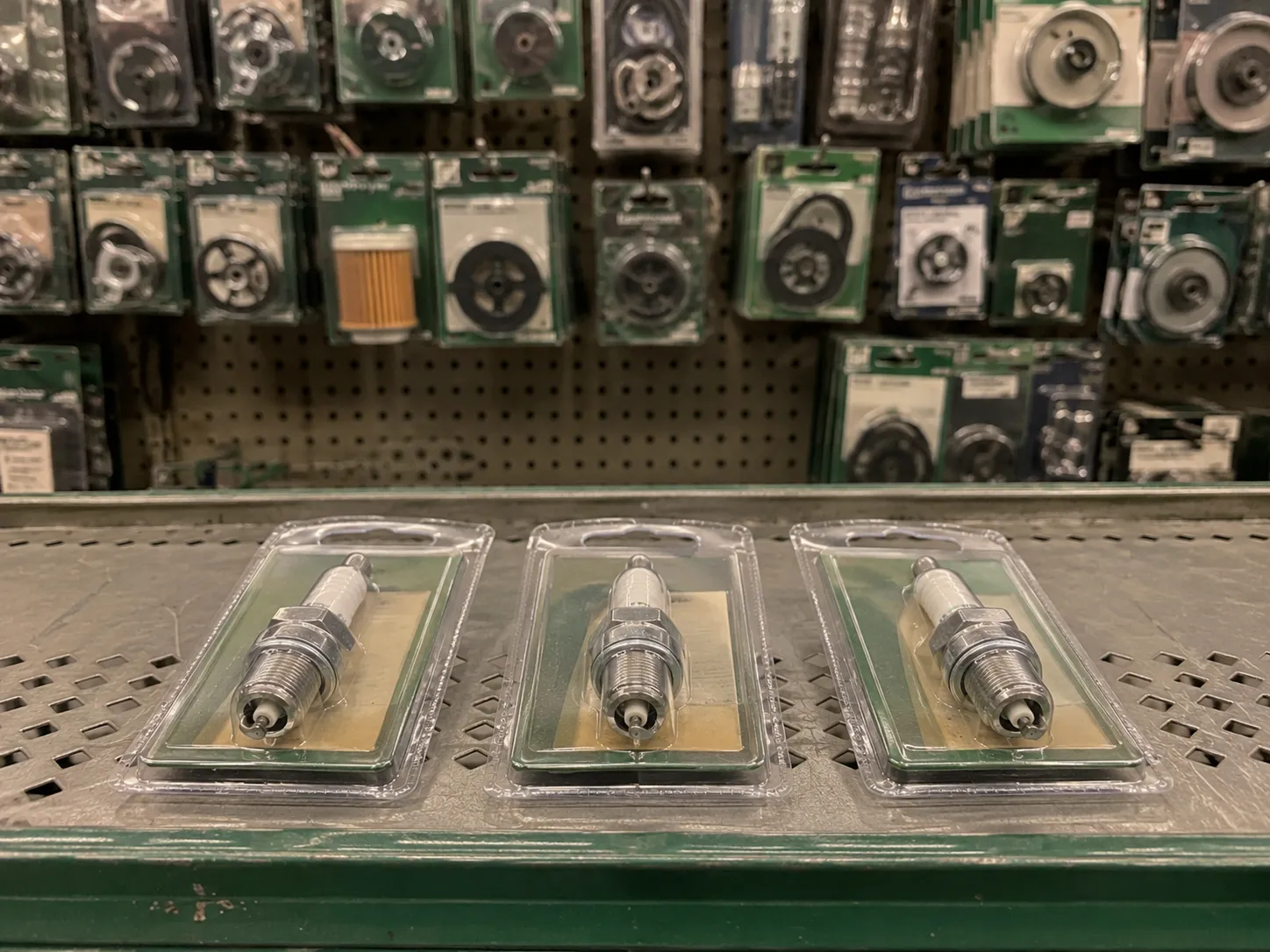 Three packaged spark plugs arranged on the front edge of a retail shelf with a blurred auto parts display in the background, illustrating in-store product availability in a small engine parts aisle.
