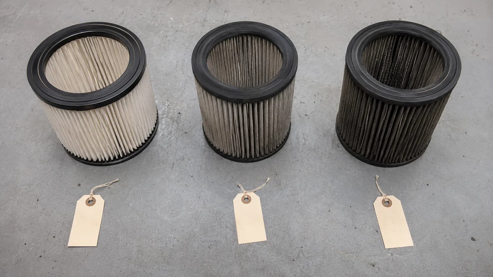 Three used cylindrical lawn mower paper air filters arranged side by side on a workbench, showing light, medium, and heavy dust loading after 128 hours of seasonal use.
