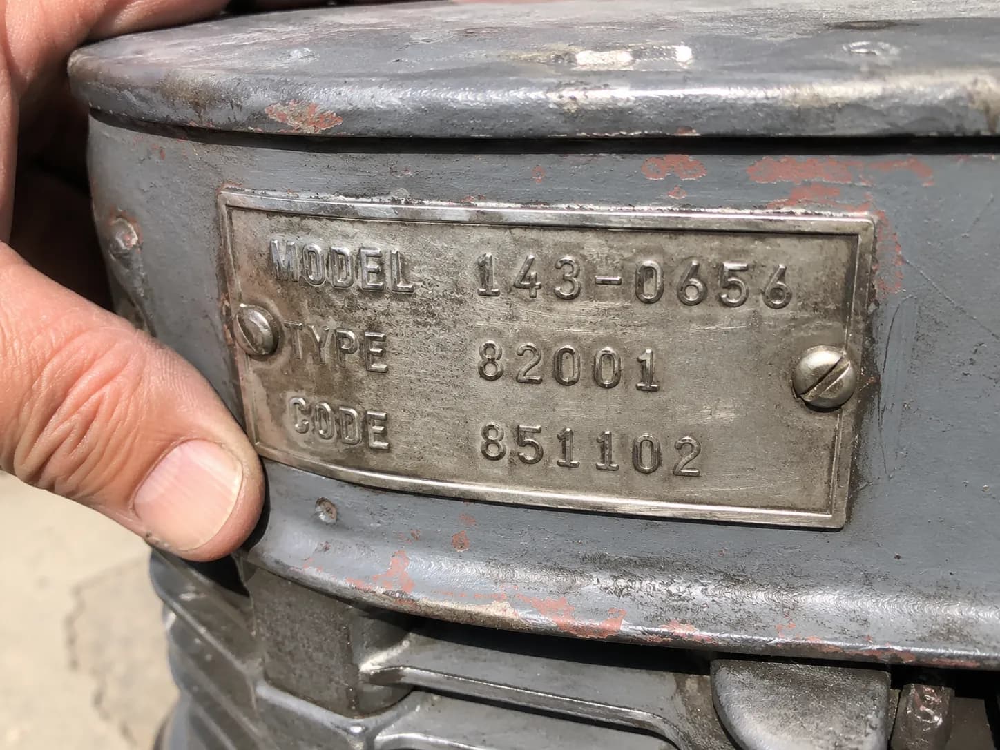 Close-up of a Toro engine identification plate showing the stamped model, type, and code numbers in daylight, with a hand steadying the engine shroud for clear, legible identification.