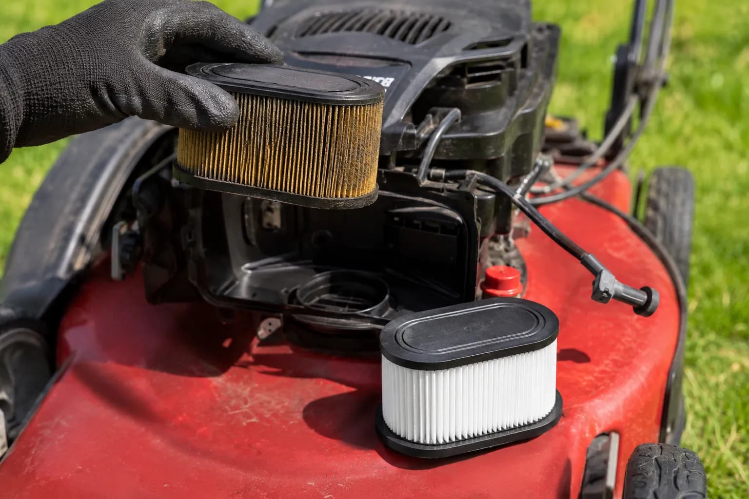 Gloved hand removing a dirty air filter from a Toro Recycler lawn mower while a clean replacement filter rests beside the open air-filter housing with the spark plug lead disconnected.