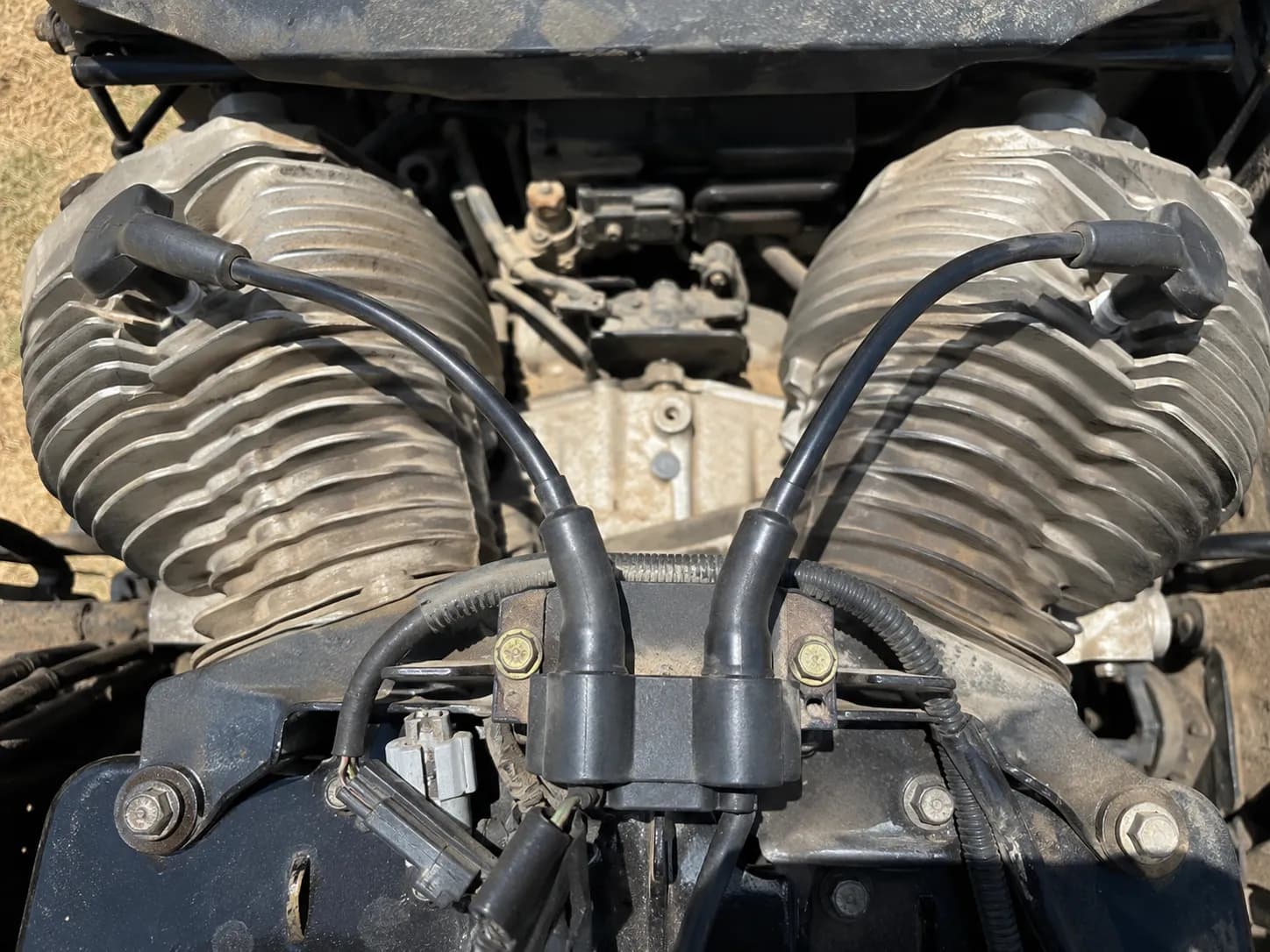 Top-down view of a twin-cylinder small engine showing an ignition coil with two spark plug leads routing in opposite directions to separate cylinder heads with dusty cooling fins visible.