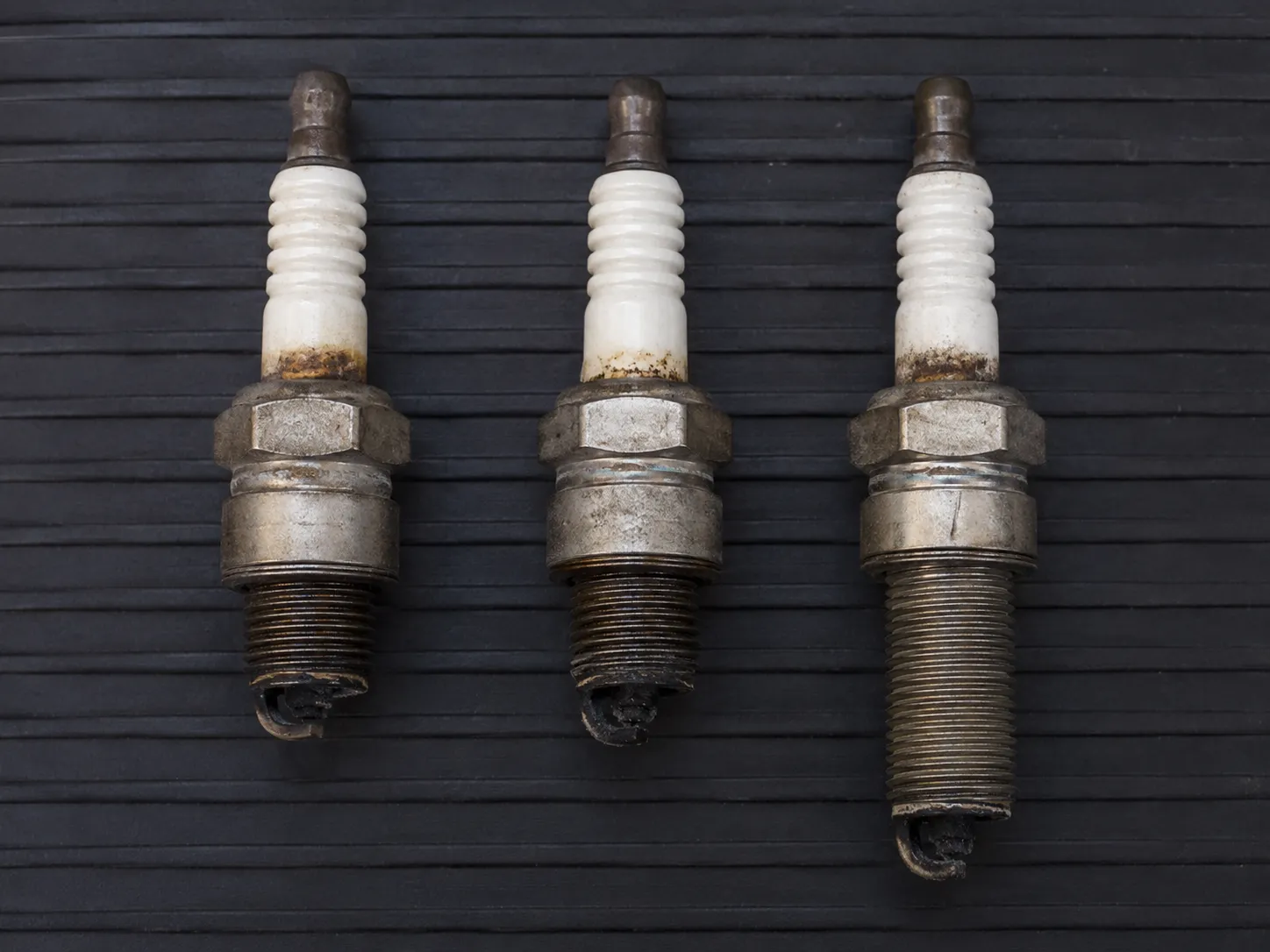 Three used Briggs lawn mower spark plugs aligned at the seating faces, comparing two short-reach plugs with one long-reach plug to show the difference in threaded body length.