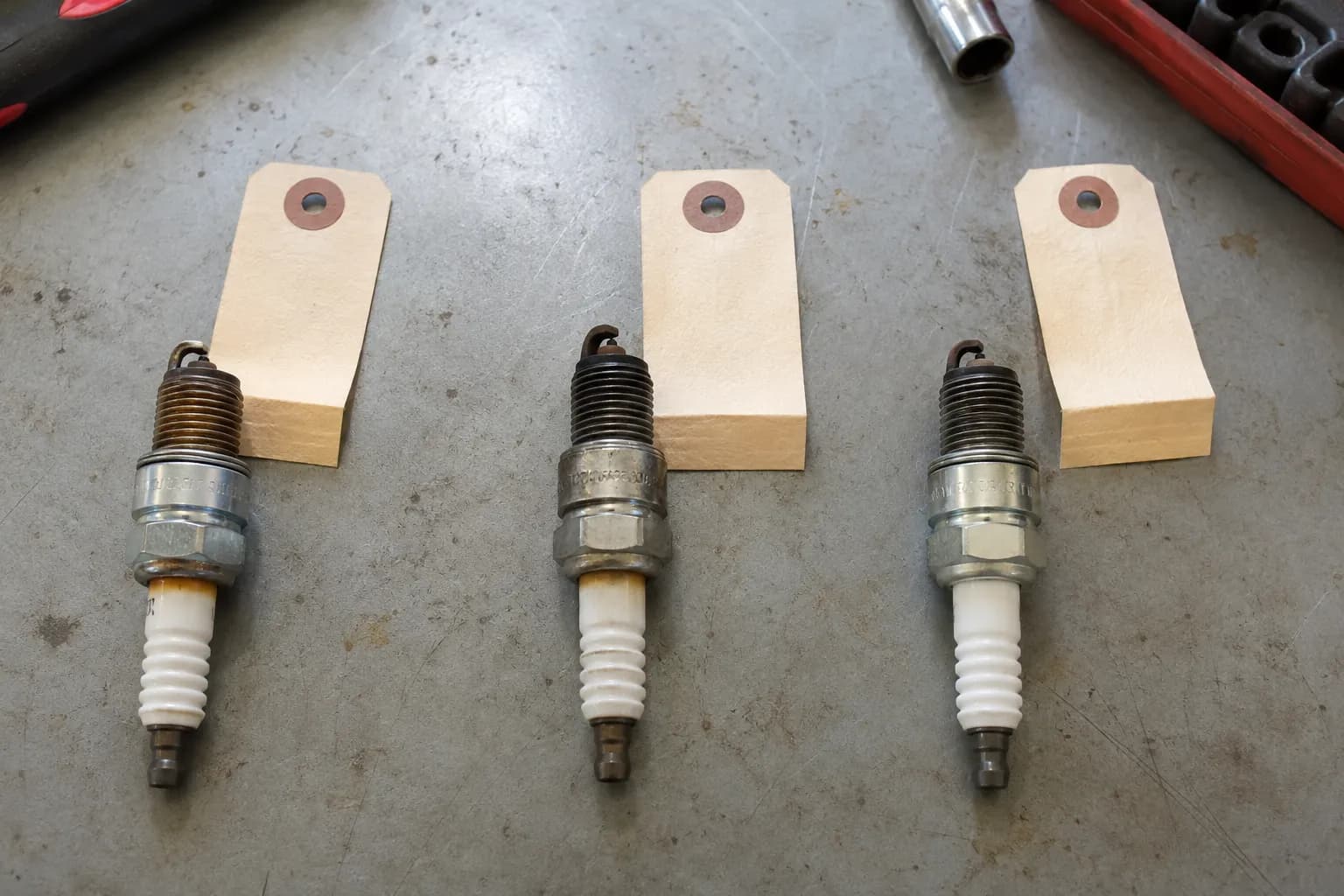 Three used Honda mower spark plugs, including two BPR5ES and one BPR6ES, arranged on a gray workbench beside paper model tags, showing different wear and tip colors.