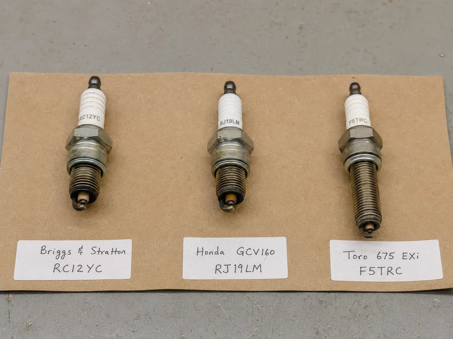 Three used lawn mower spark plugs laid on kraft paper with handwritten engine labels, comparing thread reach and electrode design on a gray workbench.