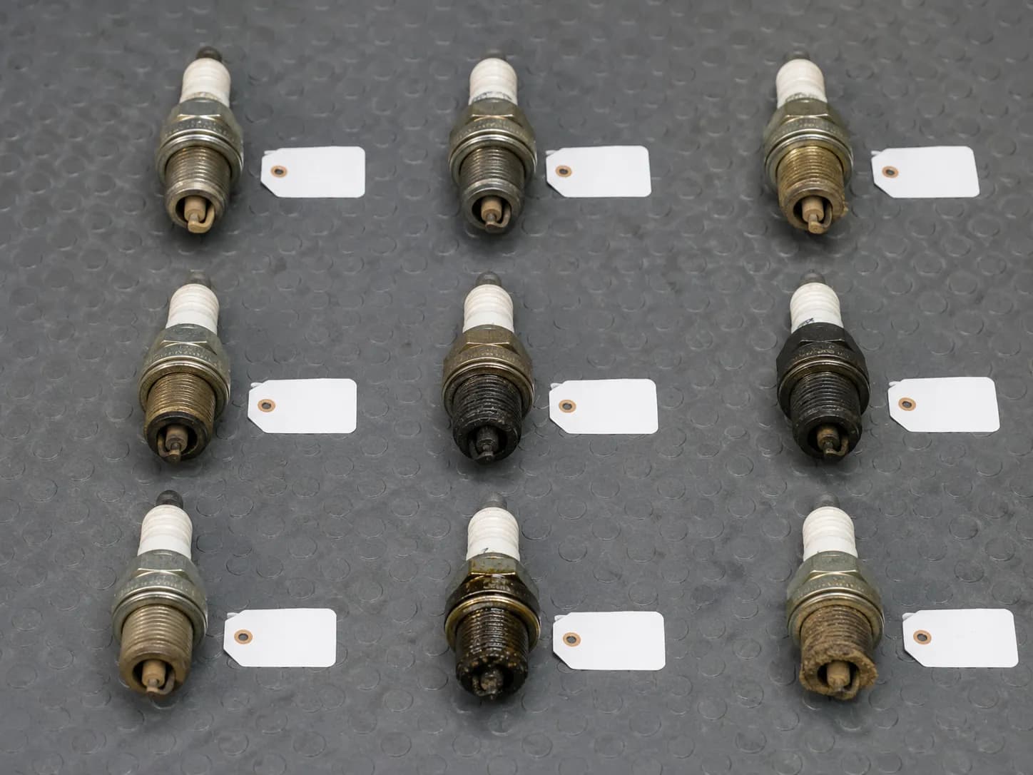 Nine used lawn mower spark plugs arranged in a 3x3 grid on a workshop mat, showing clean, sooty and oily electrode conditions with blank tags beside each for comparison after testing.