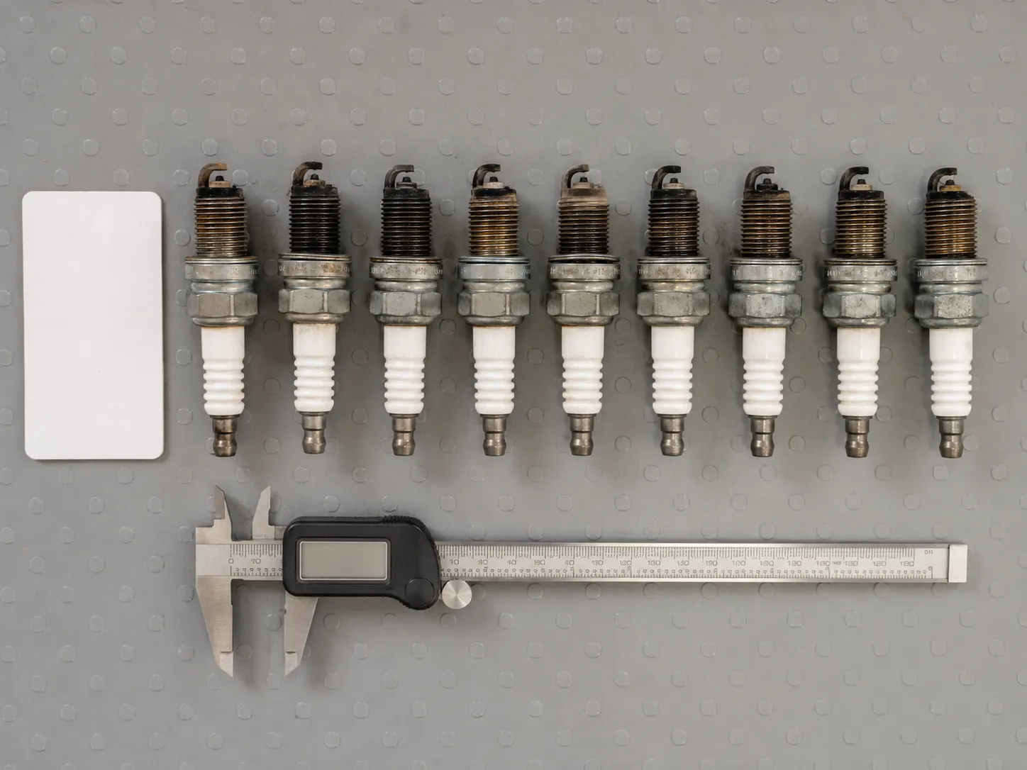 Nine used lawn mower spark plugs arranged in a numbered row on a grey rubber mat with varying electrode wear and carbon deposits, measured alongside a stainless digital caliper.