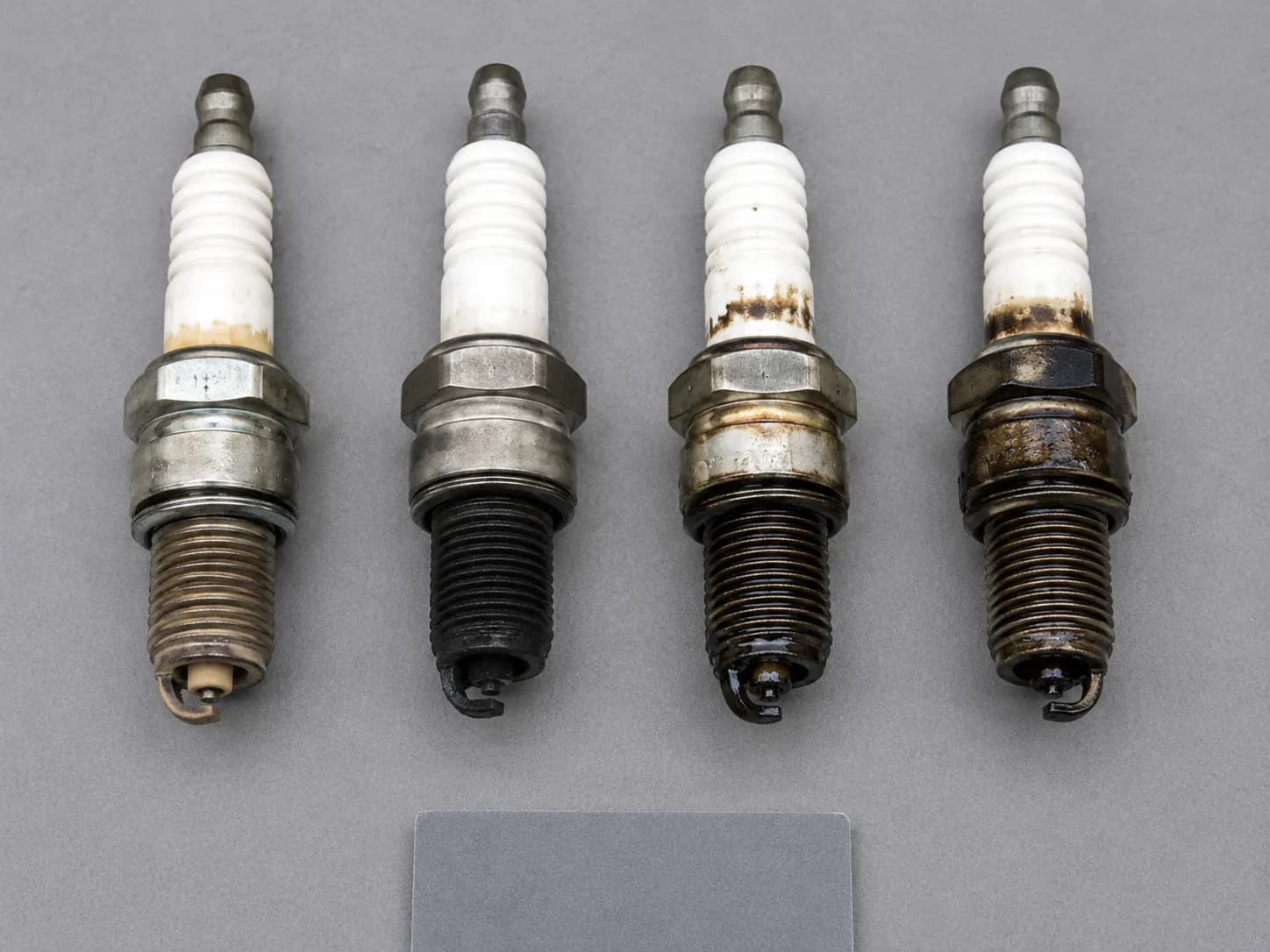 Overhead comparison of four used small-engine spark plugs under identical lighting, showing light tan, dry black soot, fuel-wet, and oil-fouled firing-end conditions side by side.