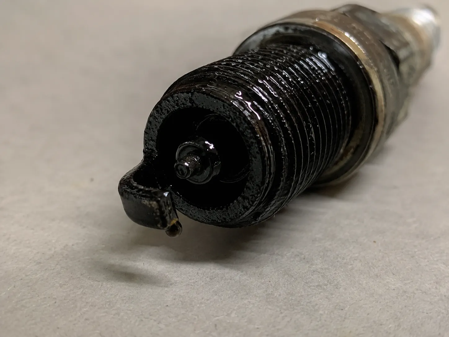 Macro photo of a used spark plug with glossy black wet oil fouling on the insulator tip and electrodes, showing heavy oil contamination from a high-hours small engine.
