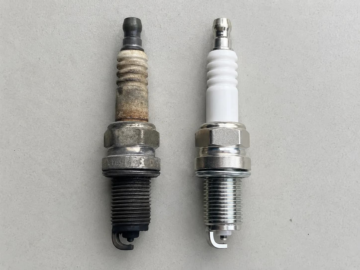 Overhead comparison of a heavily fouled used Honda lawn mower spark plug beside a clean new replacement, highlighting carbon buildup, worn electrode, and fresh threads after maintenance.