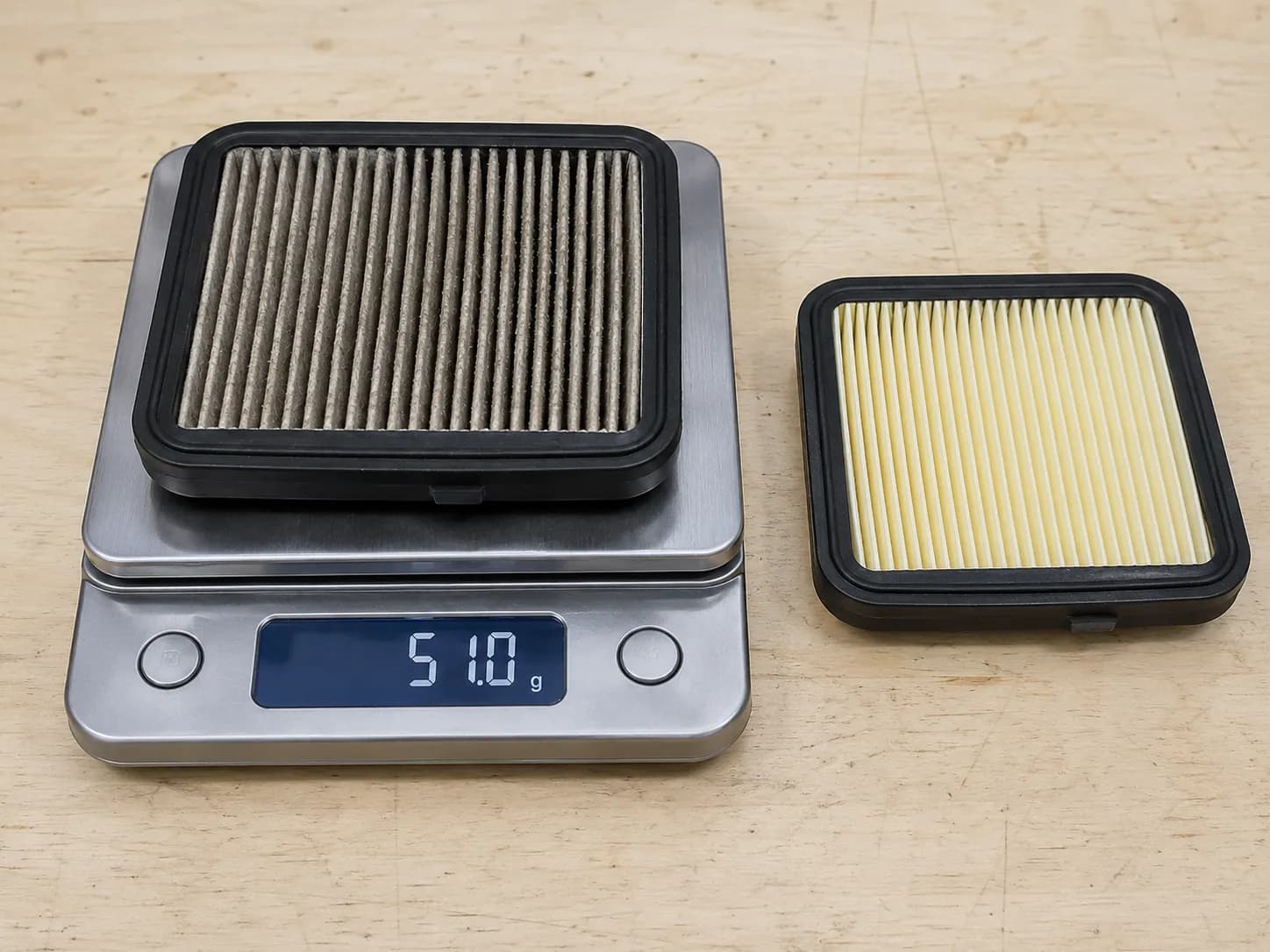 Used lawn mower pleated paper air filter on a digital kitchen scale beside a clean new replacement filter, illustrating increased weight from trapped dust after one season of use.
