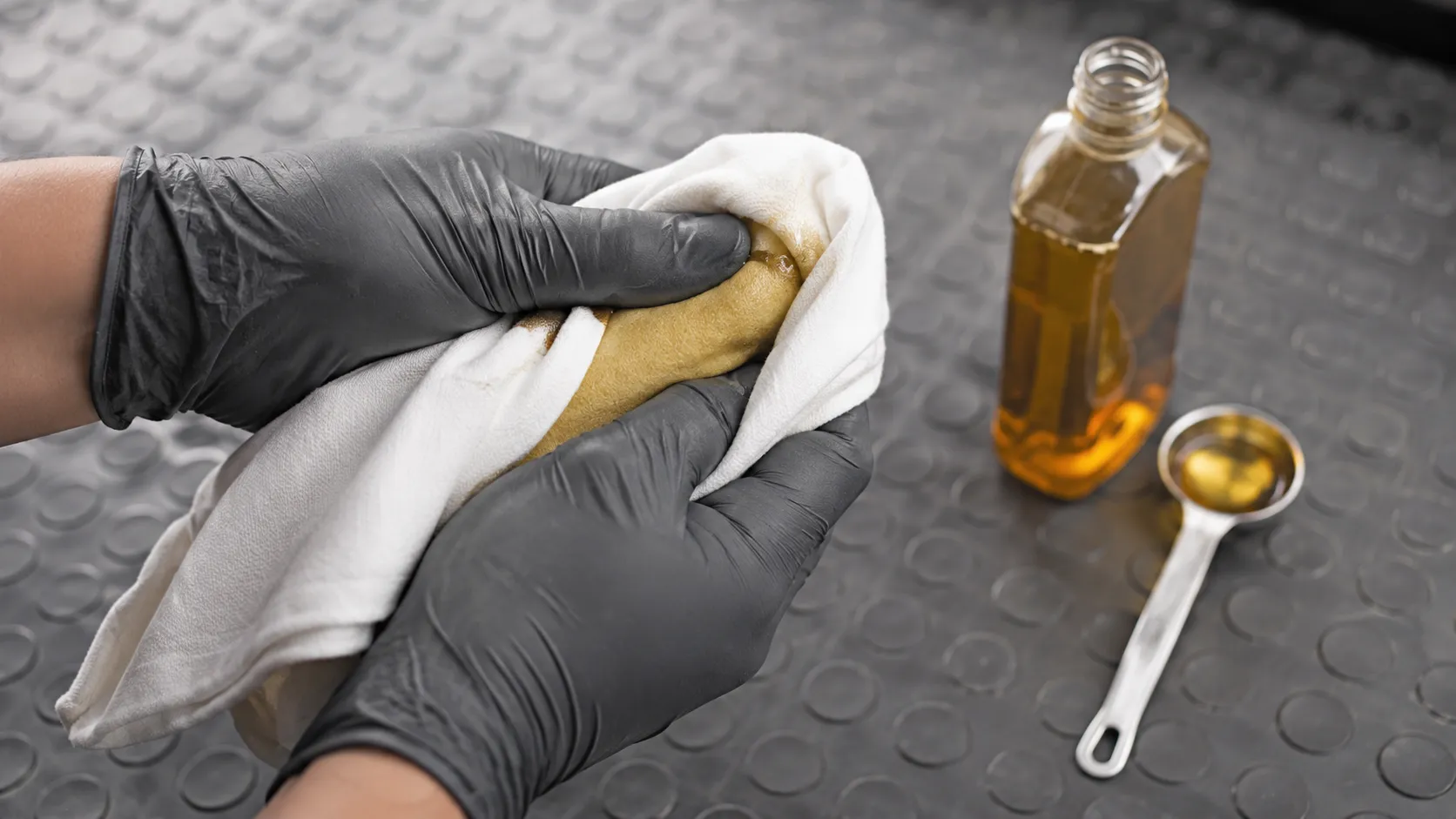 Gloved hands squeeze a freshly oiled yellow foam air filter wrapped in a white cotton cloth to remove excess oil, with an oil bottle and measuring spoon on a workshop bench.
