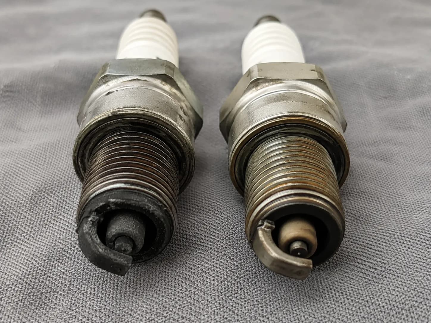 Two used YTH22V46 V-twin engine spark plugs placed side by side on a grey shop cloth, showing uneven electrode wear, carbon deposits, and measured gap differences during inspection.