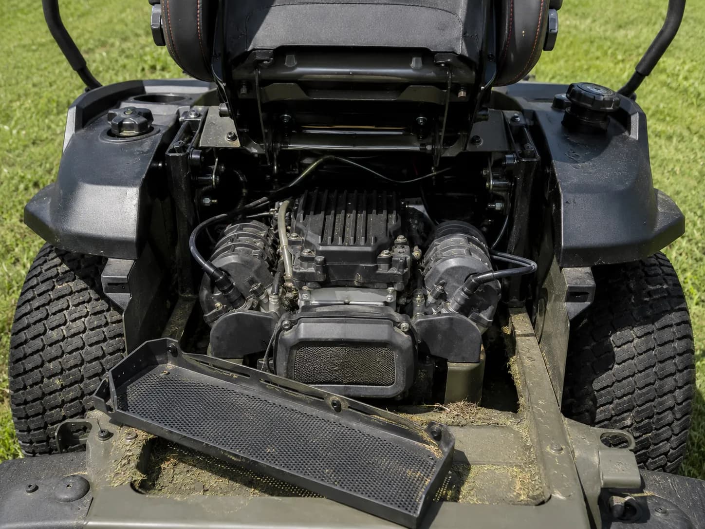 Zero-turn mower with the seat pan lifted, exposing the twin-cylinder engine below. Two spark plug leads are visible on opposite sides, with the engine guard removed for maintenance access.